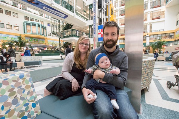 Man and woman sit together holding a baby. They are in the SickKids atrium.