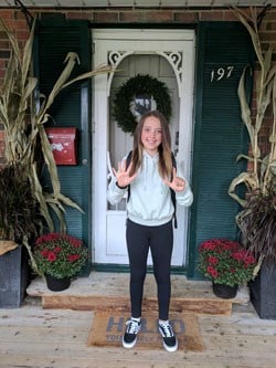 Girl stands on front porch wearing a backpack, hoodie, leggings and sneakers.