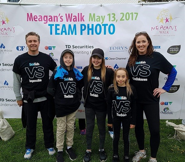 Two adults and three kids in matching shirts pose in front of a backdrop that reads: Meagan's Walk May 13, 2017 Team Photo