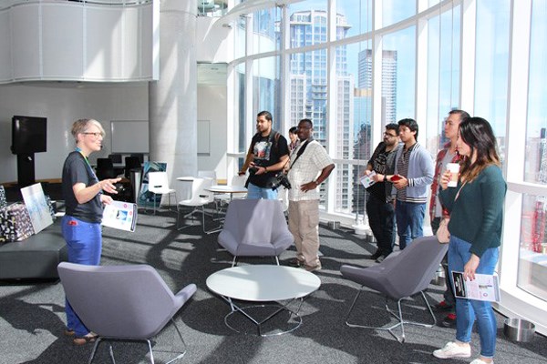 People stand in a brightly lit space with windows. One person speaks to the group.