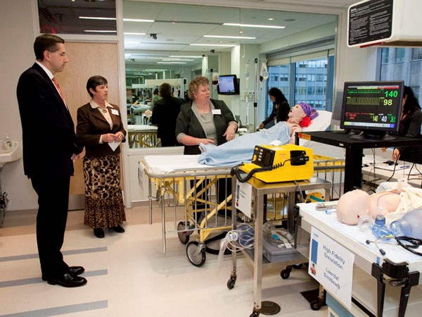 A man in a suit observes as two women present a medical simulation space including dummy on a stretcher and monitors.