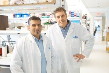 Two men stand side by side in a lab, wearing lab coats.