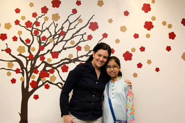 Woman has her arm around a young girl and they both face the camera. Behind them is a mural of a tree with orange and red leaves.