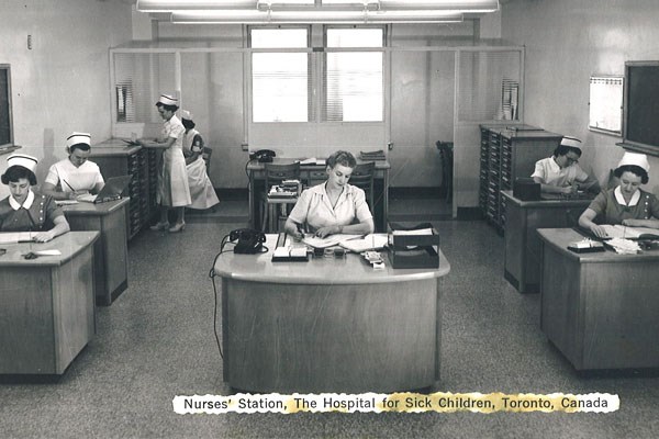Black and white photo showing five women sitting at individual desks. Some stand in the back looking at files.
