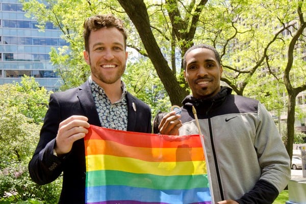Two men stand side by side, one holds a rainbow flag.