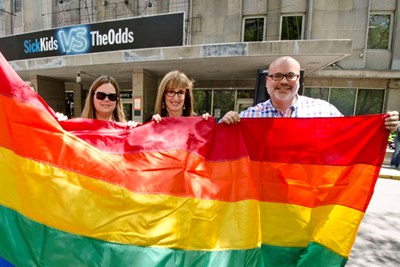 Three people stand together behind a very large rainbow flag.