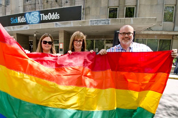Three people stand together behind a very large rainbow flag.