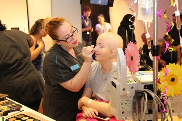 Woman applies makeup to a teenage girl who has an IV pole next to her.