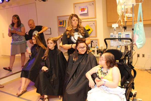Four teen girls are seated, three wear salon capes. Adults fix their hair.