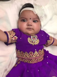 Baby lays on white sheet wearing ornate purple dress.