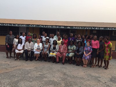 Large group pose for a photo with one row seated and one row standing behind. They are in front of a building with a sign that reads "College of Nurses & Midwives."