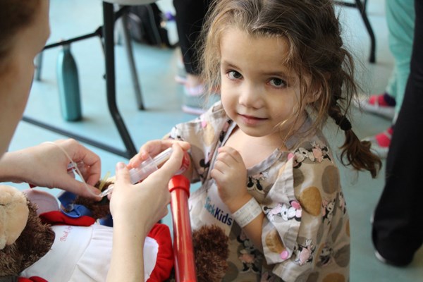 Girl holds thermometer against a teddy bear with an adult's help.