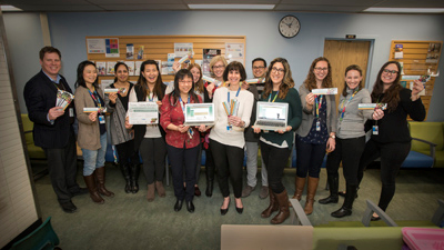 A group of fifteen adults stand together for a group photo, some holding bookmarks and others holding laptops with the screen facing the camera.