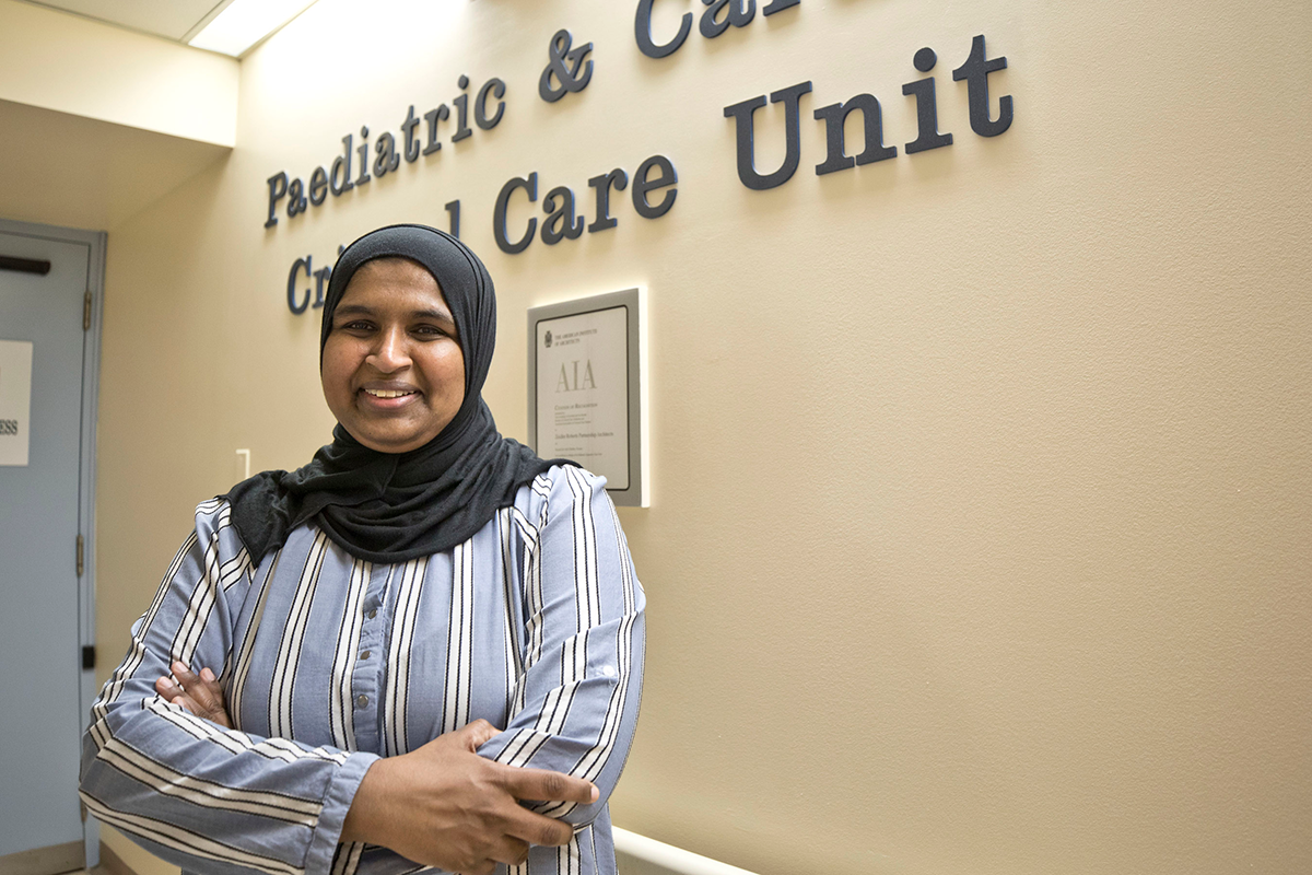 Asma stands smiling in front of the Paediatric Critical Care Unit sign in a SickKids hospital hallway