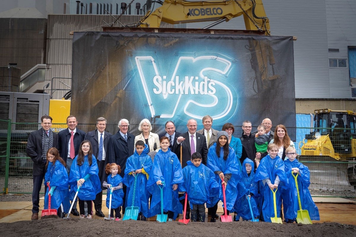 Group of children holding shovels wearing blue ponchos. A line of adults dressed formally stand behind them.