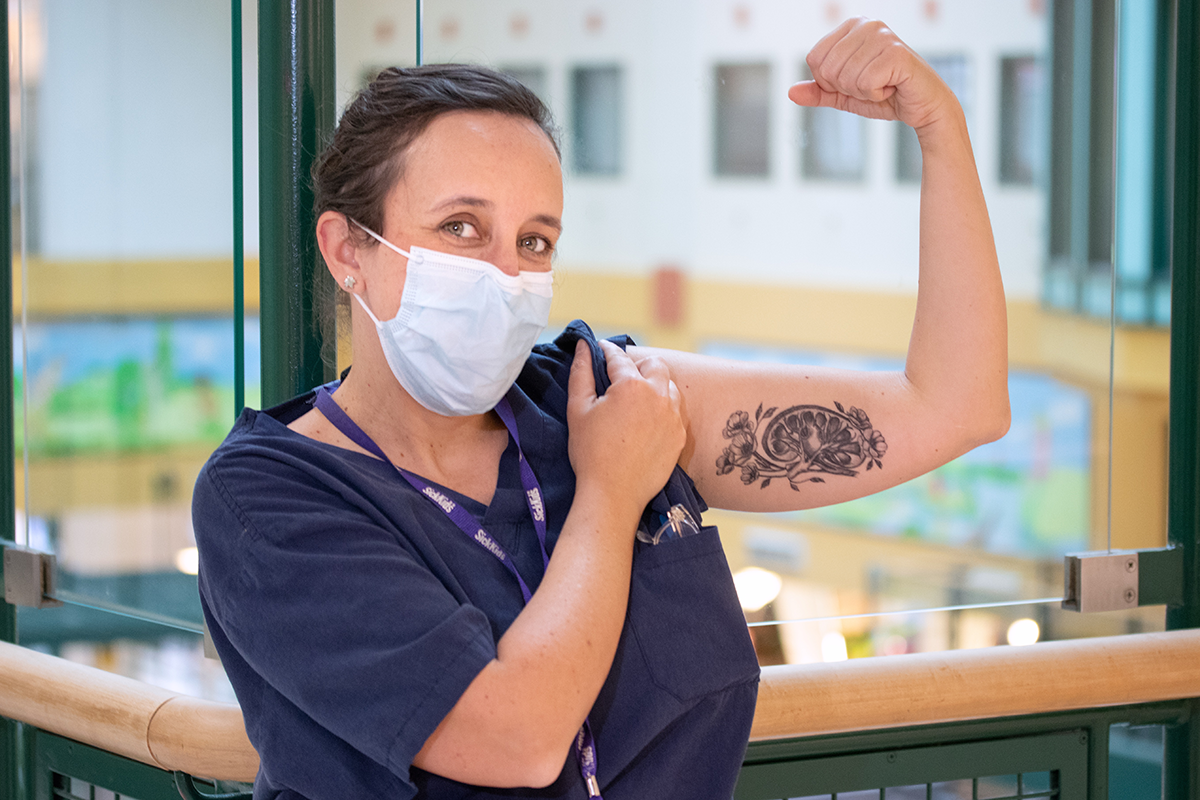 Karen Milford wearing scrubs and a face mask, flexes her arm to reveal an artistic bicep tattoo of a brain and flowers