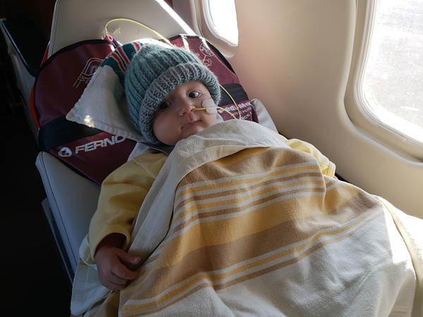 A small baby with a NG tube in his nose sits bundled in blankets in an airplane seat