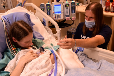 Woman in hospital bed holding a baby. Man wearing mask sites next to her. Many machines in the background.