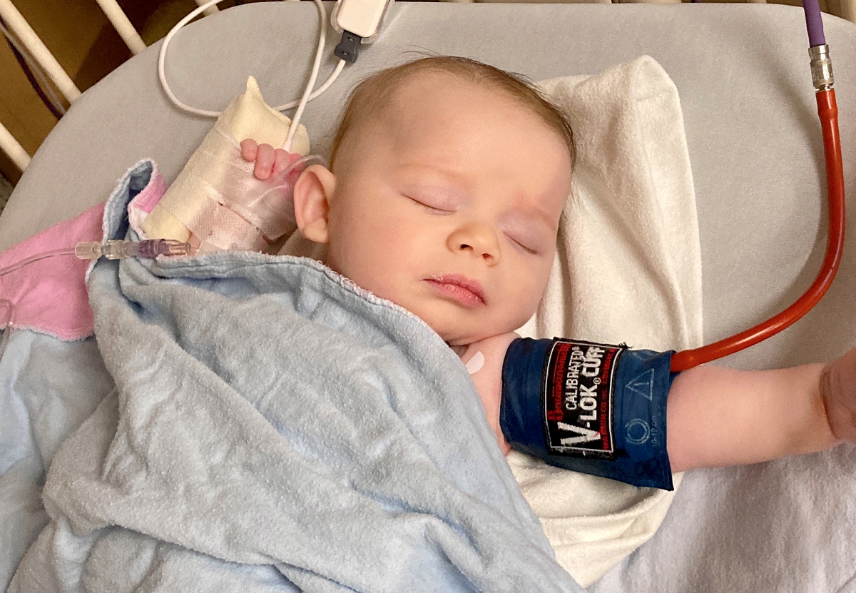 Baby lays on back with eyes closed in a hospital bed with a blood pressure cuff around his arm.