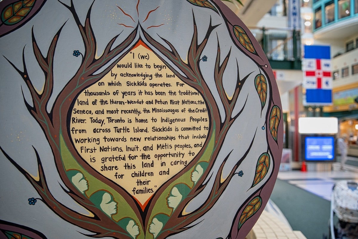 A wooden plaque painted with tree branches, leaves, flowers and human faces. At the centre is the SickKids Land Acknowledgement statement, included in the image caption.