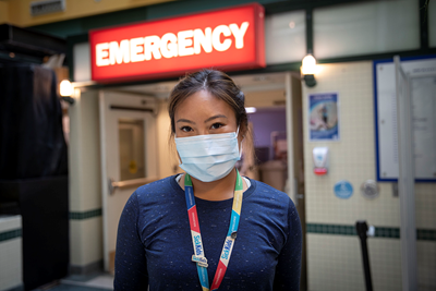 Nurse Anjelica Guytingco stands in front of a red sign that reads "emergency" in white font, wearing a mask and a SickKids lanyard. 