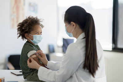 Toddler wearing a mask has a bandage put on his arm.