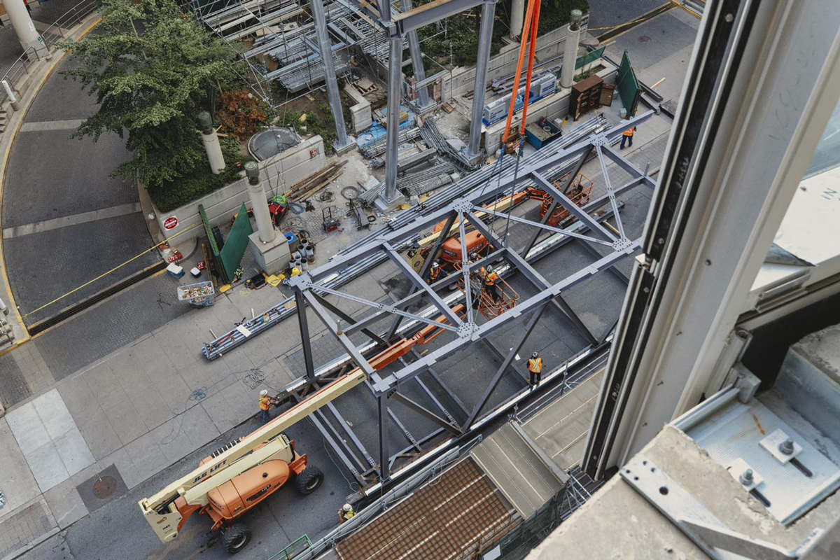 An overhead view of a city street where construction workers are assembling large steel beams into a rectangular structure