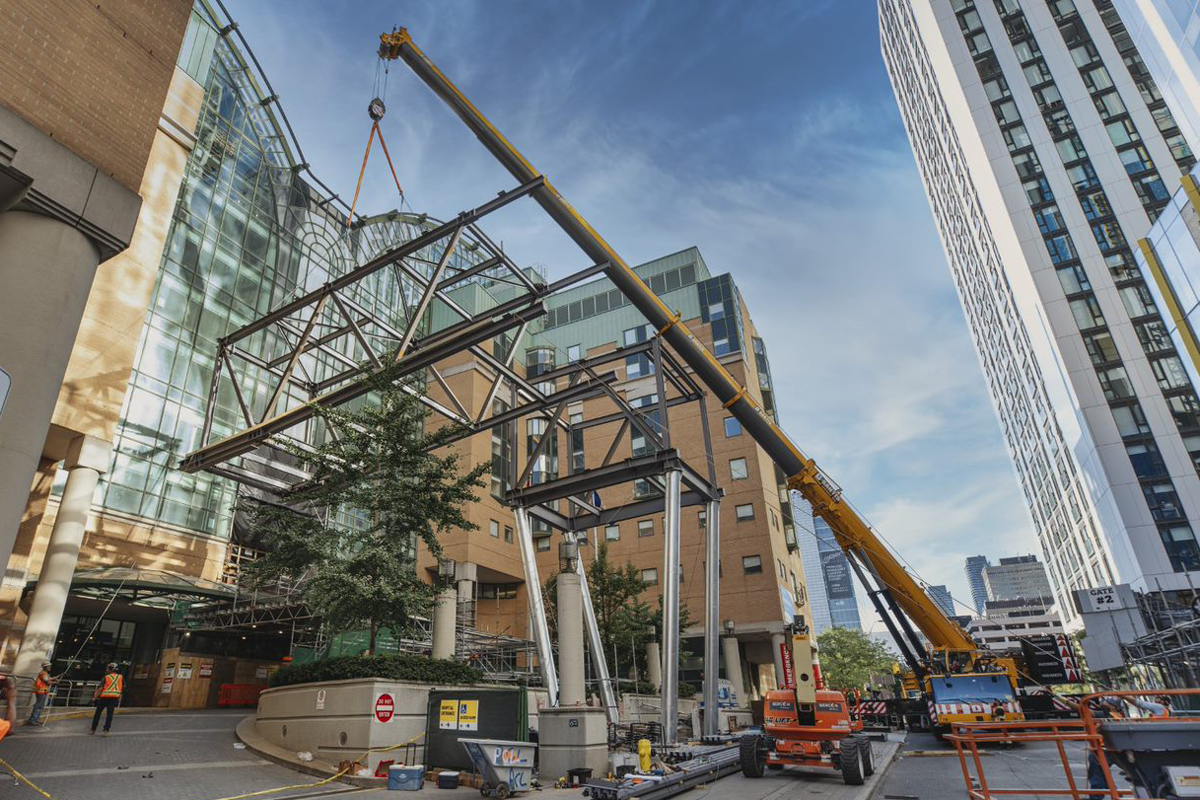 A crane on a city street lifts a large rectangular structure made of steal beams 