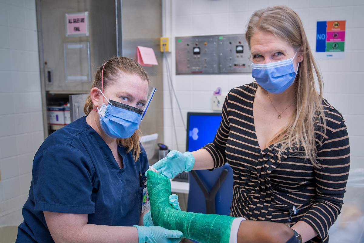 Two women wearing masks stand together working on a leg cast.