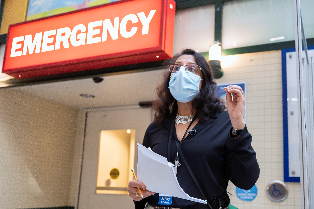 Person stands in front of Emergency department sign, wearing a mask and holding papers.