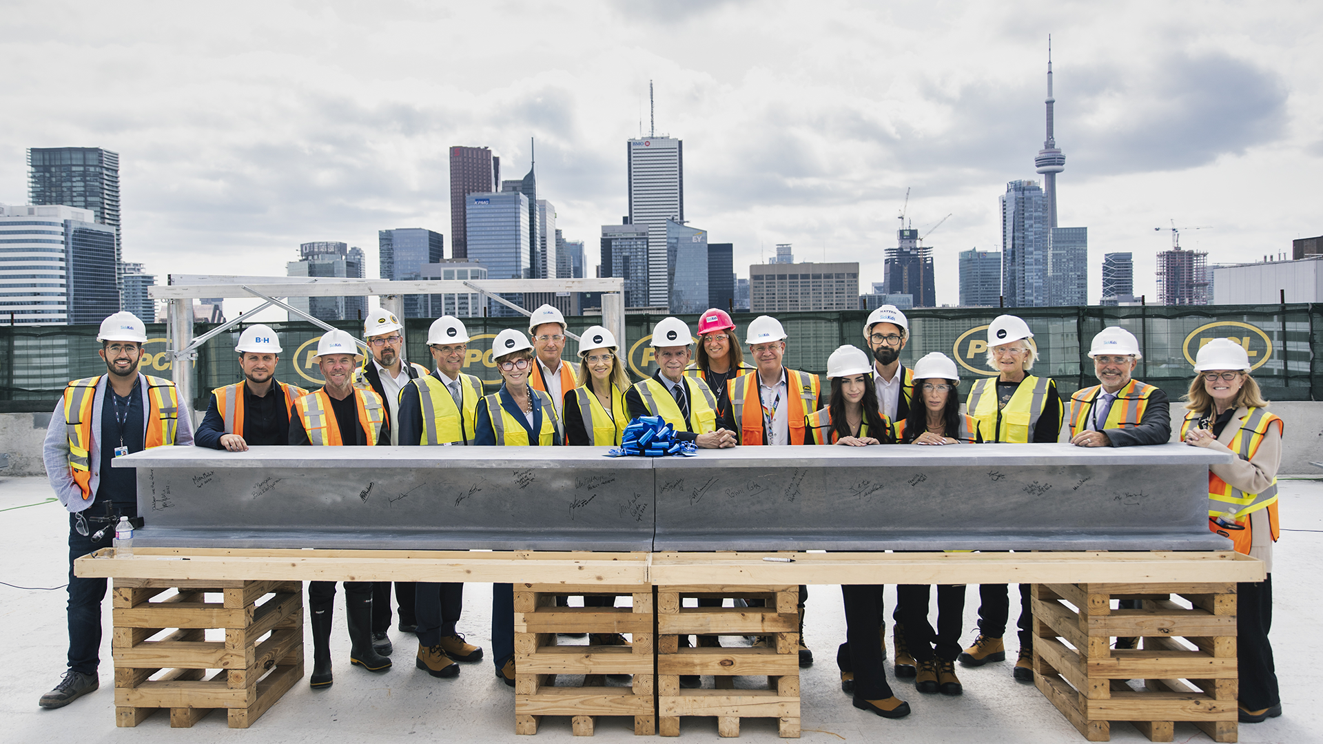 A large group of men and women wearing construction gear stand together with a large steel beam covered in signatures. The Toronto skyline is visible behind them