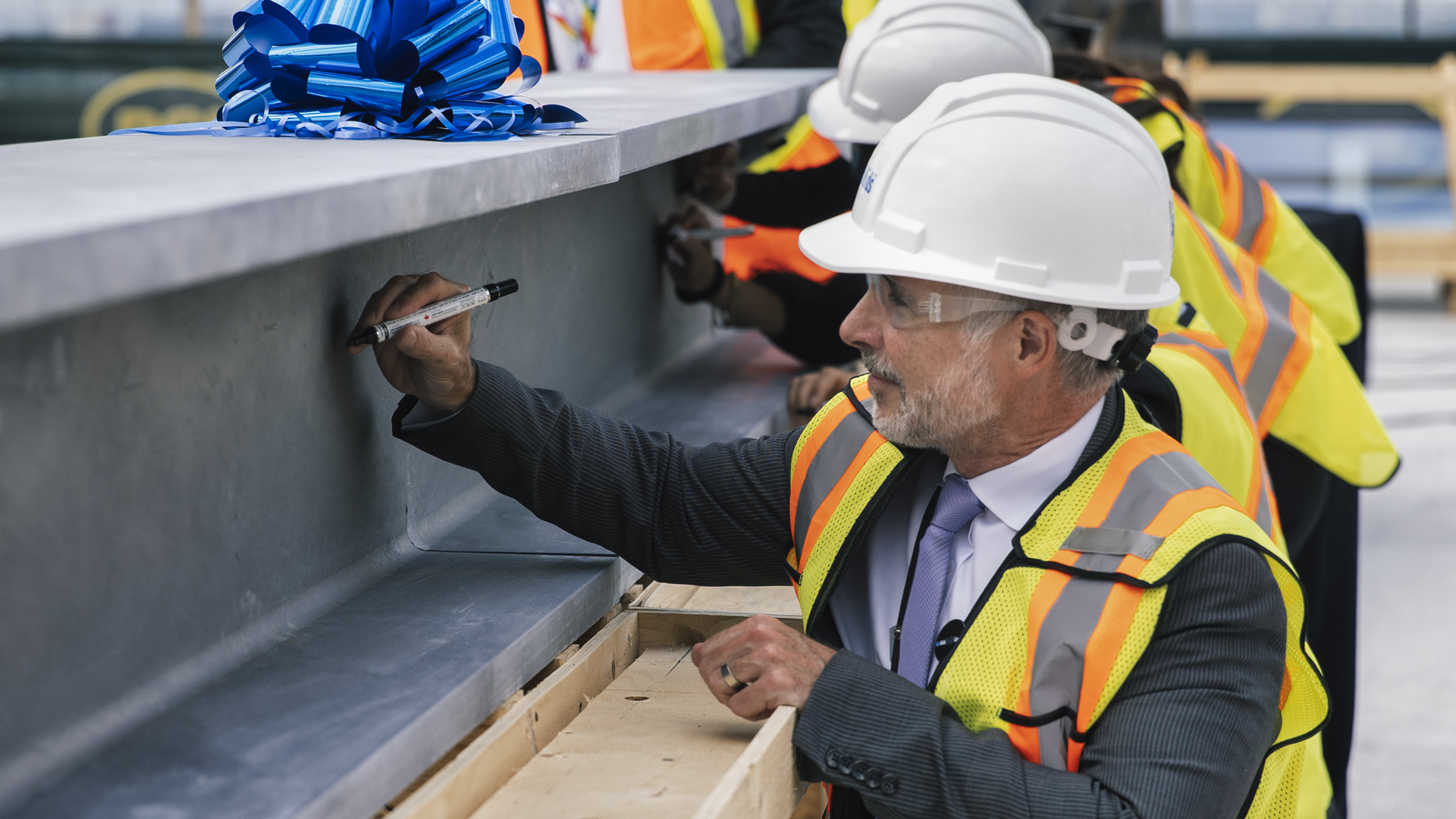 A man wearing a hard hat, glasses and a construction vest kneels down to sign a large steel beam using a permanent marker
