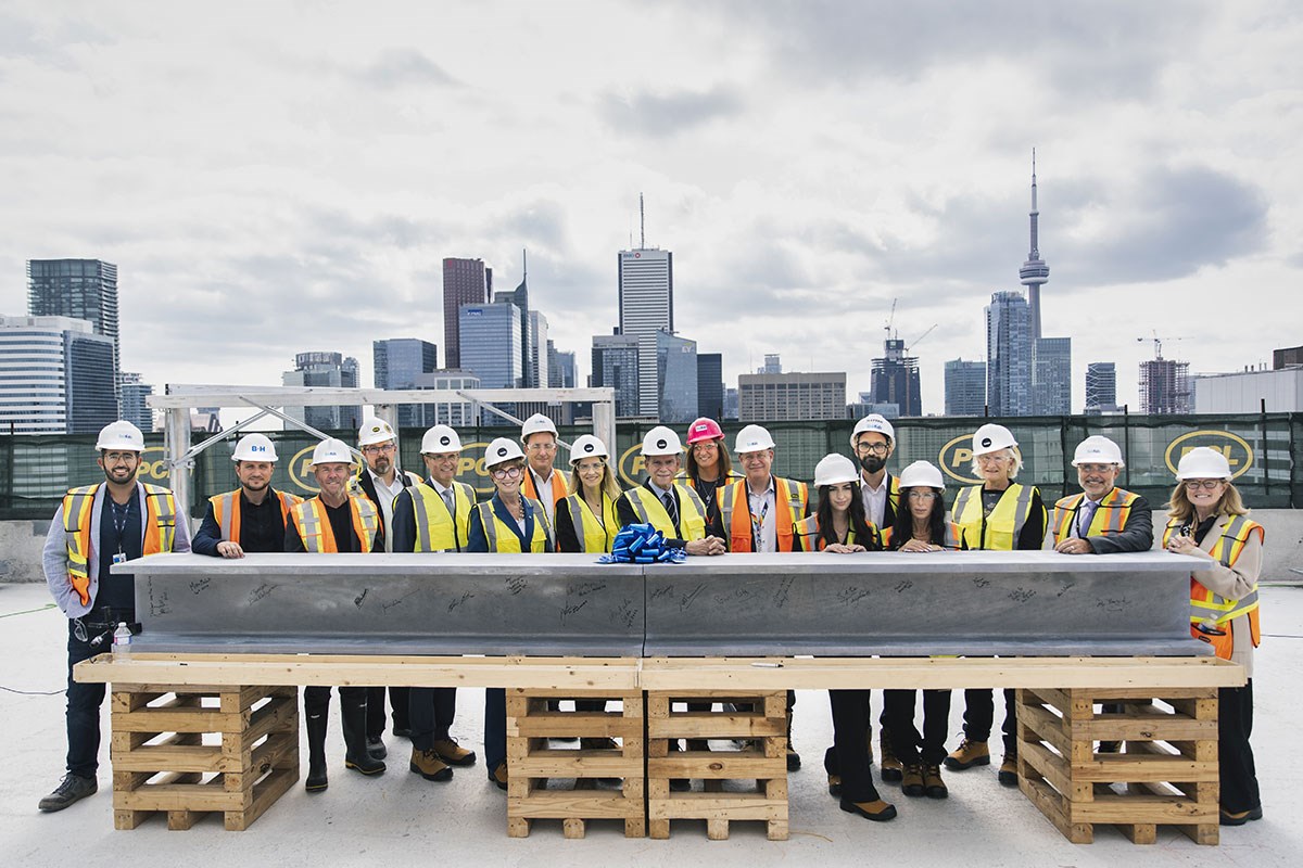 A group of people wearing construction gear gather around a large steel beam on the roof of a building, with the Toronto skyline in the background