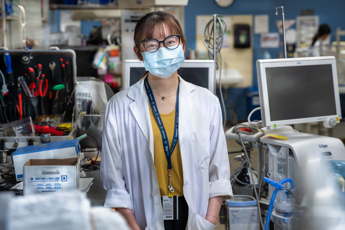 Woman wearing glasses, a mask and a lab coat stands amidst many large medical devices.