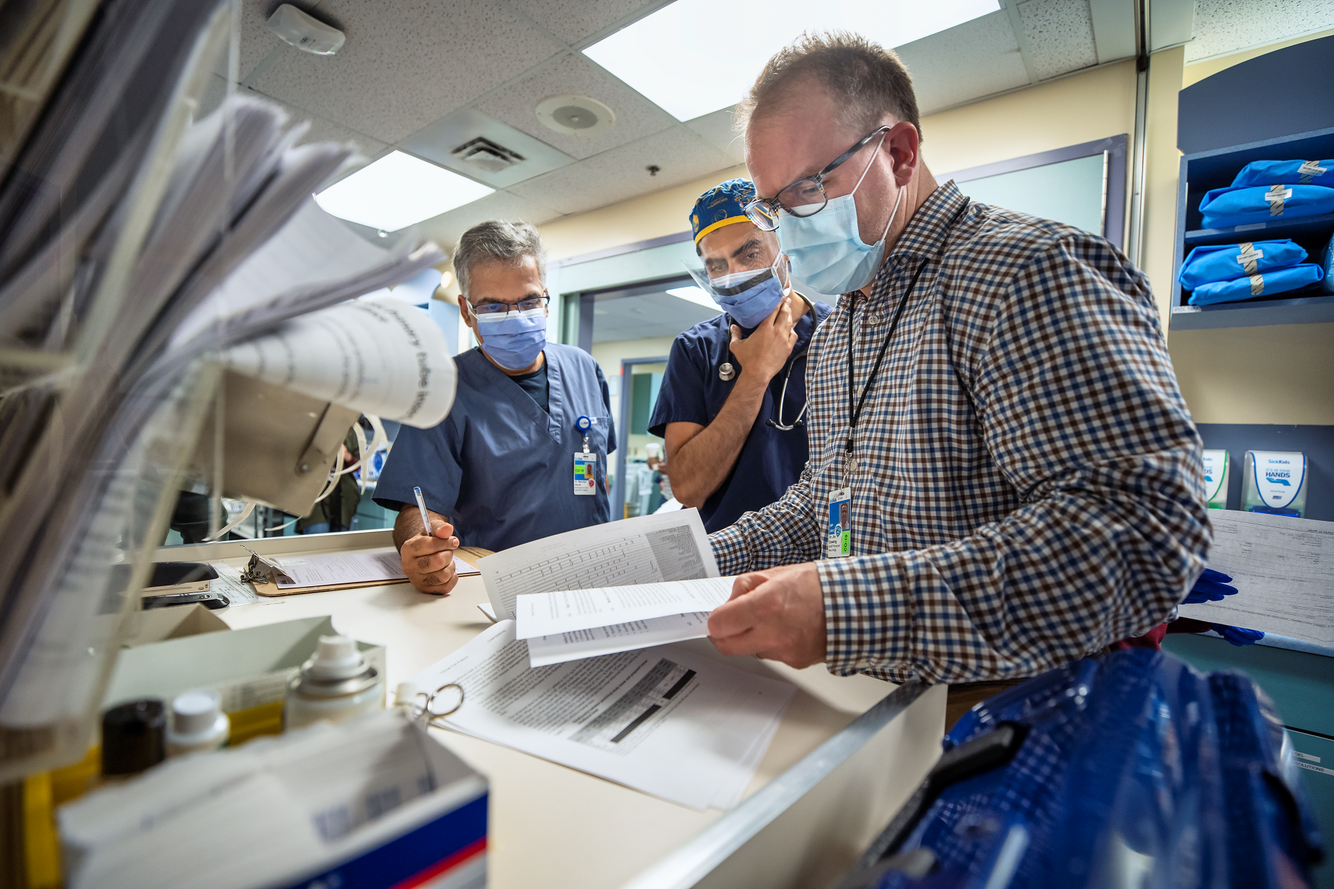 Three people stand together reviewing a document. They wear hospital masks and eye protection.