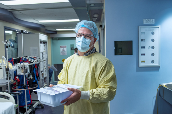 Man wearing yellow scrubs, surgical mask, hairnet and glasses. He holds a box and stands in a room filled with hospital equipment.