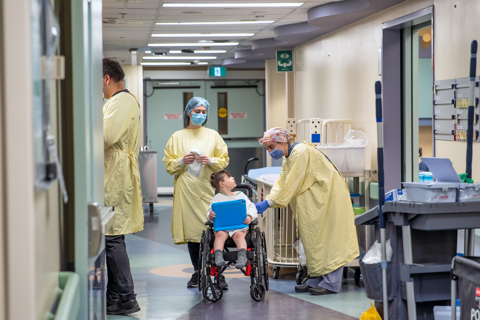 Child sits in a wheelchair holding a tablet. Three adults in medical gowns, caps and masks are present. One leans over and speaks to the child.