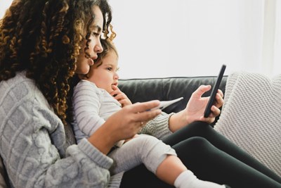 A person sits on the couch with a baby on their lap, holding and looking at their phone in their hand. 