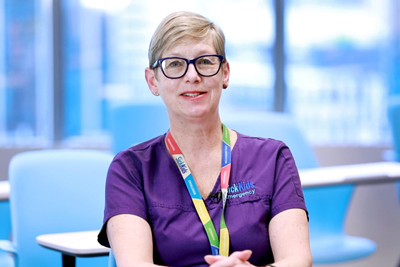 Nurse Jane Stuart-Minaret sits in a classroom with blue chairs and desks with a window in the background, wearing glasses, a SickKids lanyard and a purple shirt. 