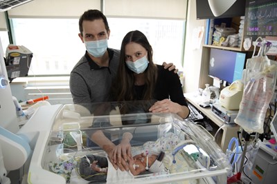 A man and woman in masks standing together with their hands resting on their baby's belly. The baby is lying in an incubator.