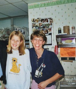 An old photo of Kristina as a child, standing with nurse, Caroline Tapper. They have their arms on each others' backs as they smile for the photo.