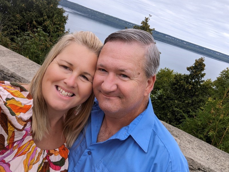 Kristina and her husband lean together for a photo in front of a lake.