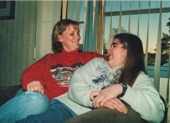 Kristina and Linda smile at each other, sitting closely on a couch in front of a window.