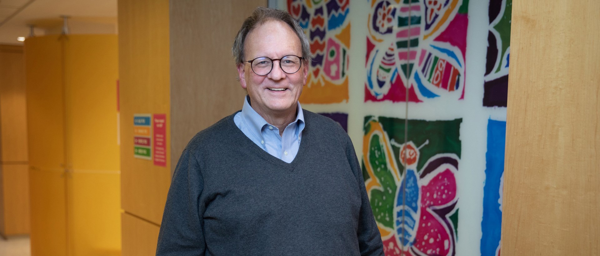 Jim Whitlock smiling and standing in a hallway in front of a wall with colourful butterfly paintings by children.