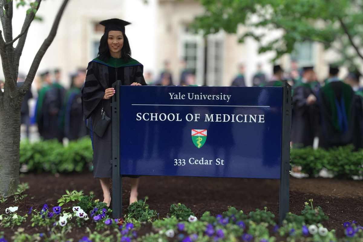 Dr. Jennifer Quon in a grad cap and regalia, standing by a sign that reads, "Yale University School of Medicine".