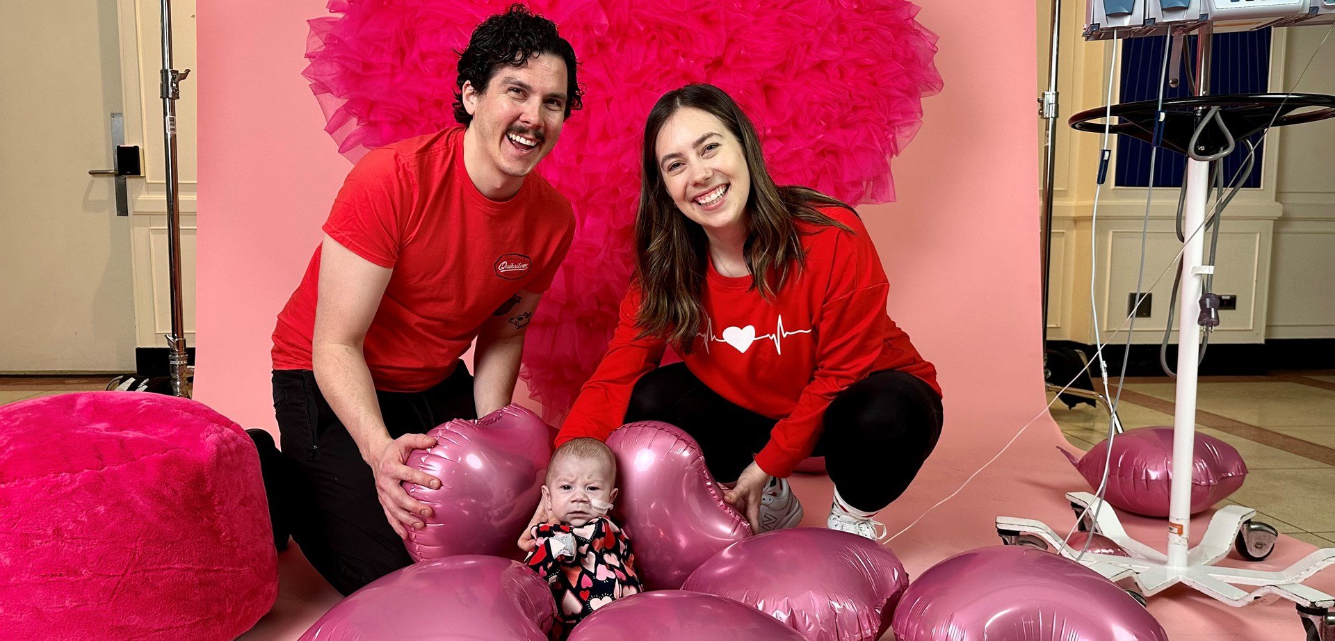 A man and a woman dressed in red T-shirts posing with their baby in front of a heart backdrop and pink heart balloons on the floor.