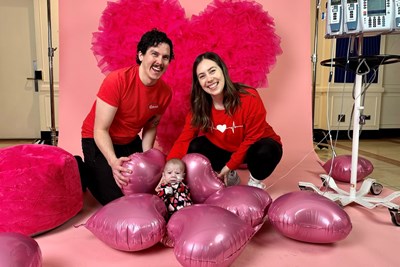 A man and a woman dressed in red T-shirts posing with their baby in front of a heart backdrop and pink heart balloons on the floor.