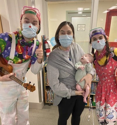 Kori Axford with her baby Milo in her arms as she poses for a photo with two therapeutic clowns.