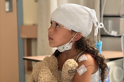 A girl sitting on a hospital bed while holding a teddy bear on her lap.