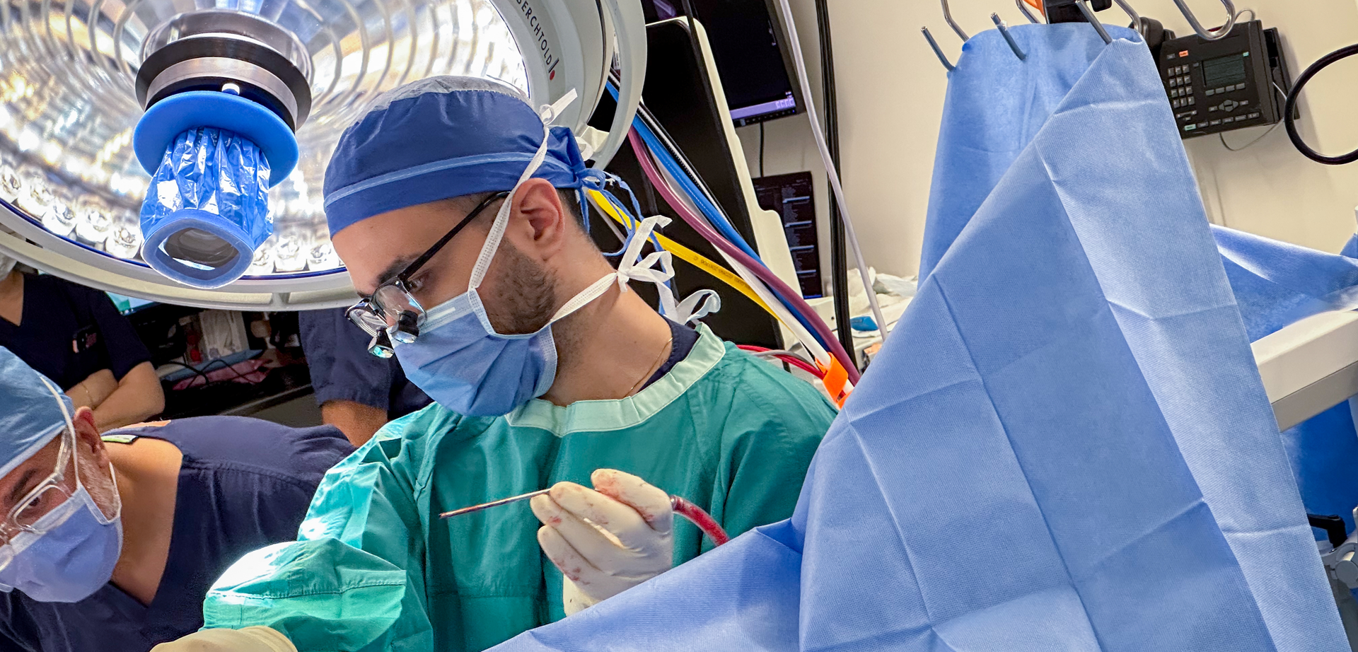 A man wearing a surgical cap, mask, glasses and robe. He is operating on an unpictured patient and is holding a surgical instrument.